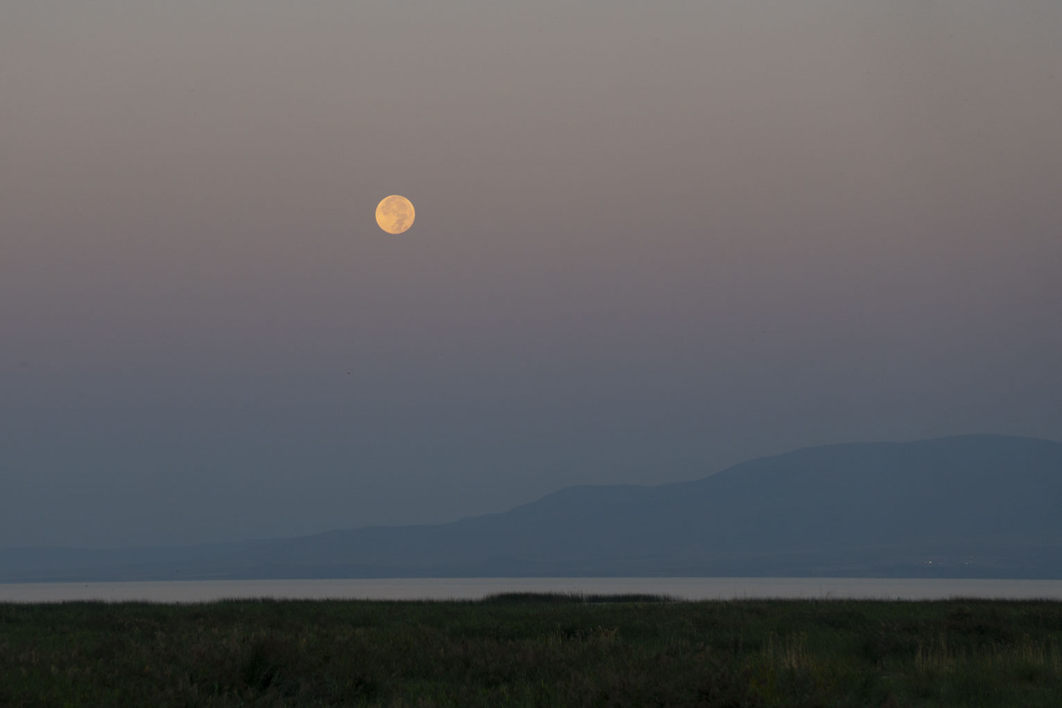 The full moon sets in dawn sky with a view of the lake and mountains beyond